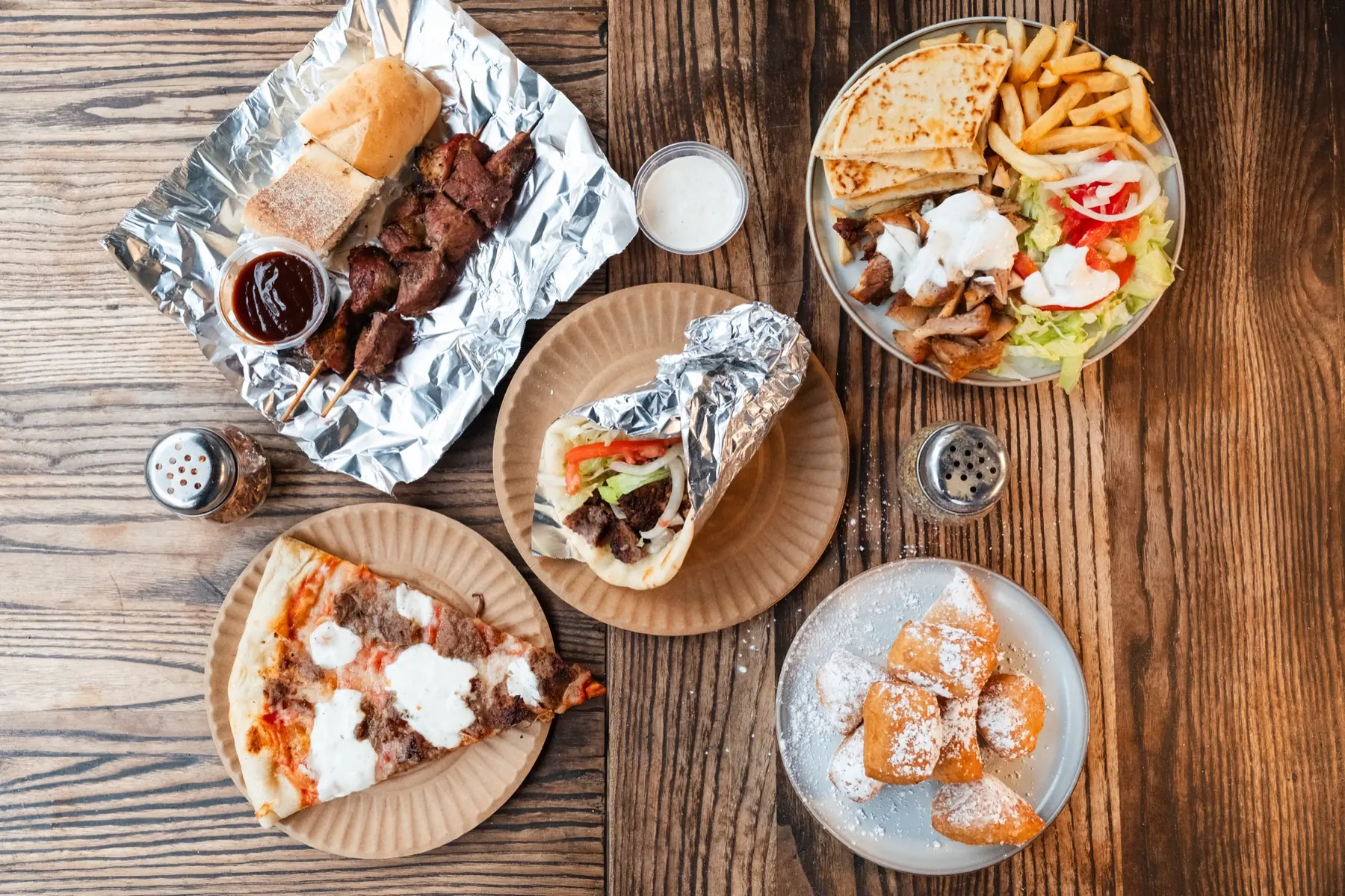 A Gyro King spread: gyro, kebab sticks, platter, gyro pizza slice and zeppoli on a wooden table